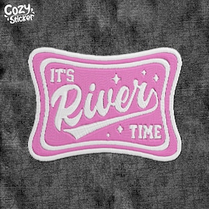 Puede incluir: Un parche bordado rosa y blanco con las palabras "IT'S River TIME" en una fuente estilizada. El parche tiene un borde decorativo y está sobre un fondo de mezclilla gris oscuro. El logotipo de Cozy Sticker está en la esquina superior izquierda.