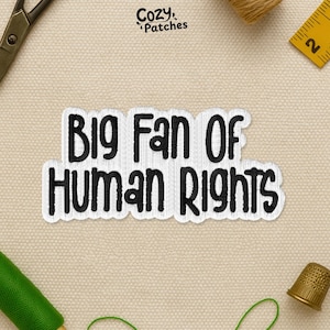 May include: Embroidered patch with the text "Big Fan Of Human Rights" in black on a white background. The patch is surrounded by sewing supplies, including scissors, thread, and a thimble. The patch is from Cozy Patches.