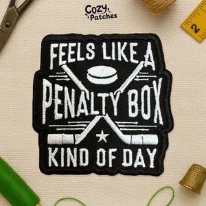 May include: Black and white embroidered patch with the text "FEELS LIKE A PENALTY BOX KIND OF DAY". The patch features crossed hockey sticks, a hockey puck, and a star. The patch is rectangular with a black border.