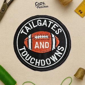 May include: Round black patch with white text that reads "TAILGATES AND TOUCHDOWNS". The patch features a football in the center, colored orange and white. The patch is surrounded by a white border. The brand name "Cozy Patches" is at the top.