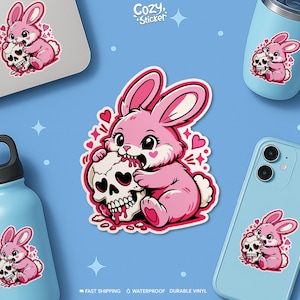 May include: Pink bunny sticker with a skull, surrounded by hearts. The bunny is biting the skull, with a cute and slightly edgy design. The sticker is on a blue background, with the text "Cozy Sticker".