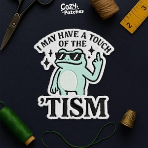May include: Embroidered patch featuring a light blue frog wearing sunglasses and giving the middle finger. The text reads "I MAY HAVE A TOUCH OF THE 'TISM". The patch is surrounded by white stitching and stars, with the brand name "Cozy Patches" at the top.