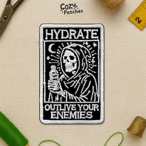 May include: Black and white embroidered patch with the text "HYDRATE" and "OUTLIVE YOUR ENEMIES". The patch features a grim reaper holding a water bottle. The patch is rectangular with a white border.