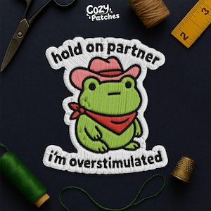May include: Embroidered patch of a green frog in a pink cowboy hat and red bandana. Text says "hold on partner" and "I'm overstimulated". The patch is white with black and green stitching. The brand name "Cozy Patches" is at the top.