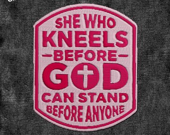 May include: A pink and white embroidered patch with the text "SHE WHO KNEELS -BEFORE- GOD CAN STAND BEFORE ANYONE" and a cross symbol. The patch has a shield shape and is set against a dark gray denim background.