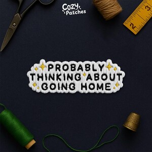 May include: Embroidered patch with the text "PROBABLY THINKING ABOUT GOING HOME" in black on a white cloud-shaped background. The patch has yellow star accents and is surrounded by sewing tools, including scissors, thread, and a thimble.