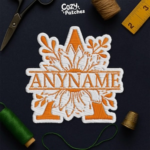 May include: Embroidered patch with the text "ANYNAME" in orange thread, surrounded by a floral design. The patch is white with orange accents and the letter "A" is visible. The patch is on a dark blue surface with sewing tools.