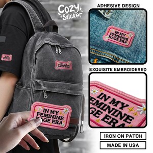 May include: Pink embroidered patch with the text "IN MY FEMININE RAGE ERA" and daisy accents. The patch is shown on a backpack and denim. The patch is an iron-on design and made in the USA.