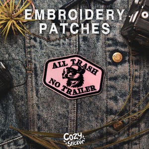 May include: Pink embroidered patch with the text "ALL TRASH NO TRAILER" and a raccoon wearing sunglasses and a hat, smoking a cigarette. The patch is on a denim jacket. The words "Embroidery Patches" are at the top.