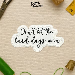 May include: A white embroidered patch with the text "Don't let the hard days win" in black script. The patch has a cloud-like shape and is surrounded by sewing supplies, including scissors, thread, and a thimble.