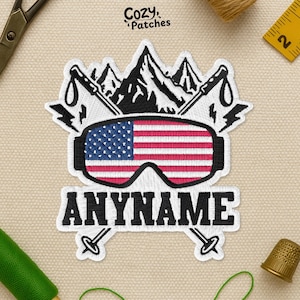 May include: Embroidered patch featuring ski goggles with the American flag design. Above the goggles are black mountains and ski poles. The text "ANYNAME" is below the goggles. The patch is white with black, red, and blue details.