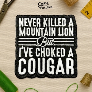 May include: Black and white embroidered patch with the text "NEVER KILLED A MOUNTAIN LION But I'VE CHOKED A COUGAR". The patch is rectangular with a black border. The brand name "Cozy Patches" is at the top.