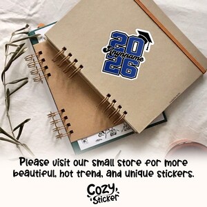 May include: A sticker with the text "2026" in blue and white, with a graduation cap graphic. The sticker also includes the text "Anynname". The sticker is on a brown notebook. The text "Cozy Sticker" is at the bottom.