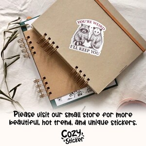 May include: Two spiral-bound notebooks with kraft paper covers. A sticker on the top notebook reads "YOU'RE WEIRD I'LL KEEP YOU" with illustrations of a raccoon and opossum. The bottom notebook has a green cover. The image also includes the text "Please visit our small store for more beautiful, hot trend, and unique stickers. Cozy Sticker."