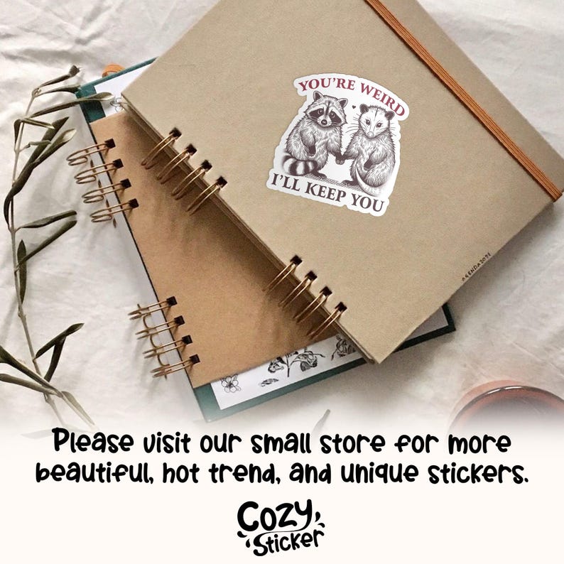 May include: Two spiral-bound notebooks with kraft paper covers. A sticker on the top notebook reads "YOU'RE WEIRD I'LL KEEP YOU" with illustrations of a raccoon and opossum. The bottom notebook has a green cover. The image also includes the text "Please visit our small store for more beautiful, hot trend, and unique stickers. Cozy Sticker."