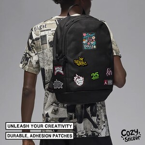 May include: Black backpack adorned with colorful patches, including text that reads "PROTECT THE DOLLS" and "WORLD CHAMPIONS." The backpack also features the text "UNLEASH YOUR CREATIVITY" and "DURABLE, ADHESION PATCHES."
