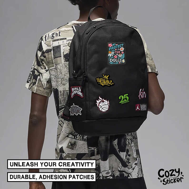 May include: Black backpack adorned with colorful patches, including text that reads "PROTECT THE DOLLS" and "WORLD CHAMPIONS." The backpack also features the text "UNLEASH YOUR CREATIVITY" and "DURABLE, ADHESION PATCHES."