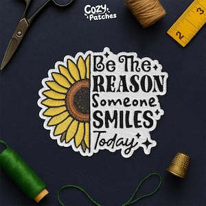 May include: Embroidered patch featuring a sunflower and the text "Be The REASON Someone SMILES Today." The patch has a white border and is set against a dark blue background. Sewing tools, including scissors, a thimble, and thread spools, are visible.