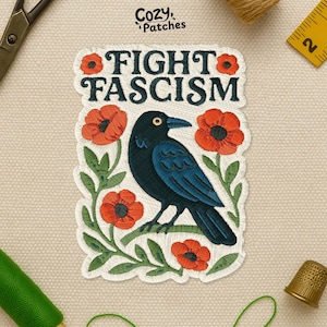 May include: Embroidered patch with the text "FIGHT FASCISM" above a dark blue crow perched on a branch with red poppies. The patch is white with a decorative border and the brand name "Cozy Patches" is in the upper left corner.