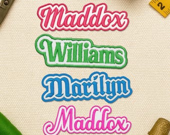 May include: Four embroidered name patches in pink, green, blue, and pink. The names are "Maddox", "Williams", "Marilyn", and "Maddox". The patches have a white border and are on a beige background. Sewing notions are in the background.
