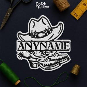 May include: Embroidered patch featuring a cowboy hat, boots, and the text "ANYNAME" in a bold font. The design is in black and white, with a detailed stitch pattern. The patch is likely for clothing or accessories.