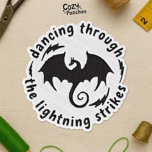 May include: Embroidered patch with a black dragon silhouette inside a white circle. The text "dancing through the lightning strikes" is embroidered around the dragon. The patch is on a beige surface with sewing tools.