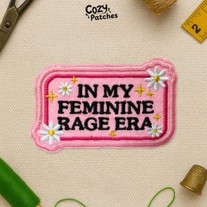 May include: A pink embroidered patch with the text "IN MY FEMININE RAGE ERA" in black. The patch is decorated with white daisies and gold star accents. The patch has a pink border and is labeled "Cozy Patches".