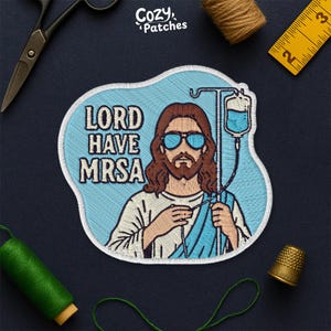 May include: Embroidered patch featuring an illustration of Jesus wearing sunglasses and holding an IV drip bag. The text "LORD HAVE MRSA" is displayed in bold letters. The patch has a light blue background and a white border.