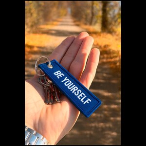 May include: A blue rectangular keychain with the words "BE YOURSELF" in white lettering. The keychain is attached to a silver keyring with several keys. The background is a blurred outdoor scene with a path and fall foliage.