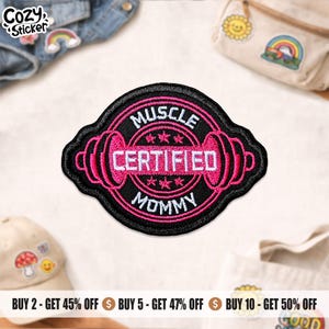May include: A black and pink embroidered patch with the words "MUSCLE CERTIFIED MOMMY" in white lettering. The patch features a barbell design and is surrounded by a black border. The patch is likely for clothing or accessories.