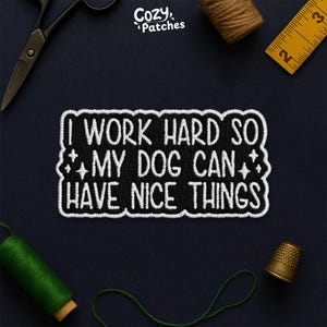 May include: Embroidered patch with the text "I WORK HARD SO MY DOG CAN HAVE NICE THINGS" in white on a black background. The patch is surrounded by a white border and decorative elements. The image also includes sewing tools.