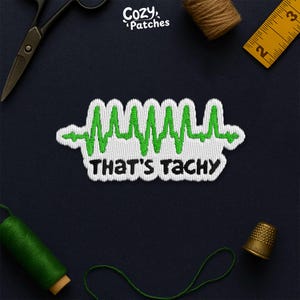 May include: White and green embroidered patch with a heartbeat design and the text "THAT'S TACHY". The patch is surrounded by sewing tools, including scissors, thread, and a measuring tape. The brand name "Cozy Patches" is in the top left corner.