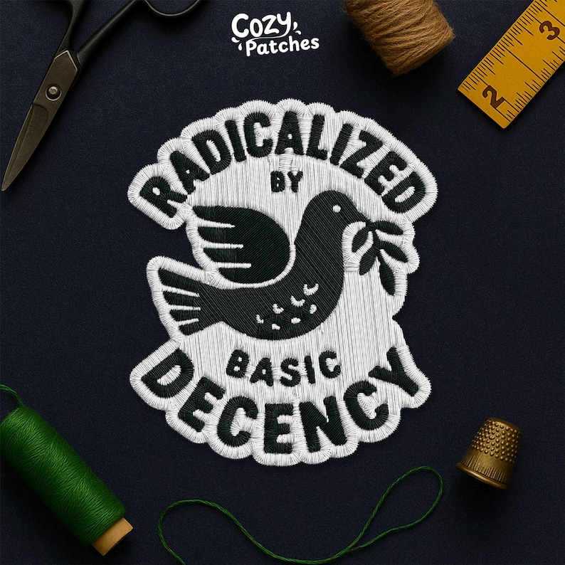 May include: Embroidered patch with the text "RADICALIZED BY BASIC DECENCY" surrounding a black dove holding an olive branch. The patch is white with black details. The brand name "Cozy Patches" is in the top right corner.