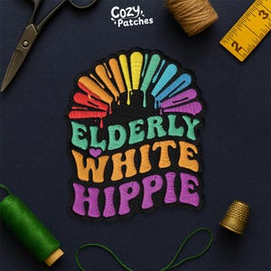 May include: Embroidered patch with the text "ELDERLY WHITE HIPPIE" in colorful, retro-style lettering. The patch features a rainbow design with dripping paint effect. The patch is on a dark blue background, with sewing tools.