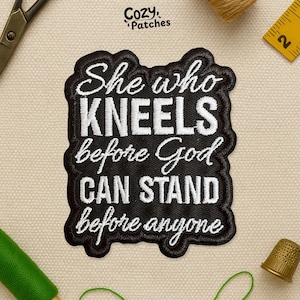 May include: Black embroidered patch with the text "She who kneels before God can stand before anyone" in white script. The patch has a black border and is on a beige fabric background.
