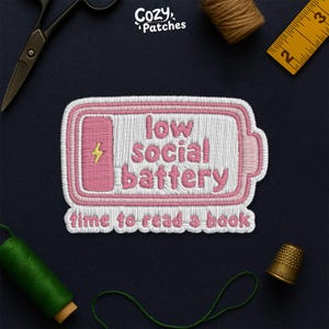 Peut inclure: Écusson brodé avec un motif de batterie rose et les mots "low social battery" et "time to read a book" en rose et blanc. L'écusson est entouré de fournitures de couture, dont des ciseaux, du fil et un dé à coudre.