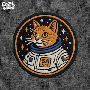 May include: Embroidered patch of an orange tabby cat wearing an astronaut suit with a blue helmet and a white spacesuit with the letters "SA" on the chest. The background is a black circle with white stars.