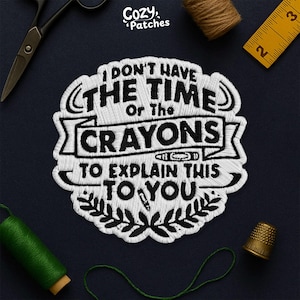 May include: White embroidered patch with the text "I DON'T HAVE THE TIME Or The CRAYONS TO EXPLAIN THIS TO YOU." The patch has black outlines and a leaf design. The patch is on a dark blue surface with sewing tools.