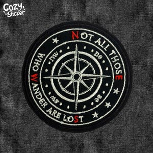 May include: A round black embroidered patch with a compass rose design. The text "NOT ALL THOSE WHO WANDER ARE LOST" is embroidered in white and red around the compass. The patch has a white border and star accents.