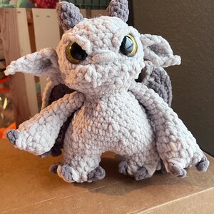 May include: A handmade crochet dragon in shades of gray and white. The dragon has large, golden eyes, small horns, and wings. The plush toy is standing on a brown surface.