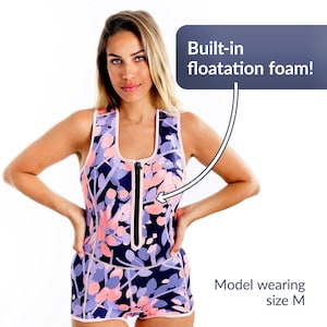 May include: A sleeveless swimsuit with a floral pattern in shades of pink, purple, and navy blue. The swimsuit has a front zipper and white trim. The text says "Built-in floatation foam!" and "Model wearing size M".