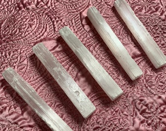 May include: Five white selenite wands on a pink quilted surface. The wands are smooth and have a natural, slightly rough texture.