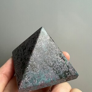 May include: A dark gray, green, and purple pyramid-shaped crystal with a rough, textured surface.