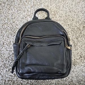 May include: Black leather backpack with a top handle and multiple zippered compartments. The backpack has a front pocket and a braided zipper pull. The zippers are gold-colored.