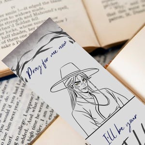May include: A bookmark featuring a black and white illustration of a character with long hair and a hat. The text "Pray for me now" is written in blue script. The bookmark is placed on top of an open book.