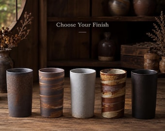May include: Five cylindrical ceramic tumblers arranged in a row, each featuring a distinct finish. The textures range from matte black and speckled brown to a light grey, a dark brown with horizontal brush strokes, and a rustic brown swirl pattern.