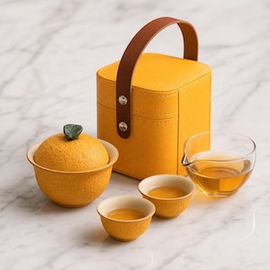 May include: A tea set featuring a yellow textured lidded bowl shaped like an orange with a green leaf handle, two small matching yellow tea cups, a clear glass pitcher filled with amber tea, and a yellow square carrying case with a brown handle.