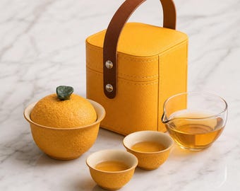 May include: A tea set featuring a yellow textured lidded bowl shaped like an orange with a green leaf handle, two small matching yellow tea cups, a clear glass pitcher filled with amber tea, and a yellow square carrying case with a brown handle.