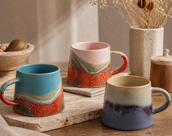 May include: Three ceramic mugs with handles, each featuring a distinct two-tone glaze. One mug is blue and red, another is pink and orange with a speckled texture, and the third is a muted beige and deep purple with a smooth, glossy finish.