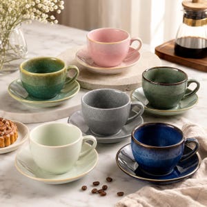 May include: A set of six ceramic teacups and saucers in shades of green, pink, grey, blue, and pale green. Each cup sits on its matching saucer. The cups are arranged on a marble surface, with coffee beans and a pastry visible.